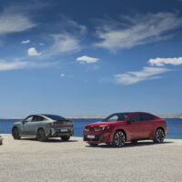 Dve verzie nového BMW iX4 na pobrežnej ceste, červená a sivá.