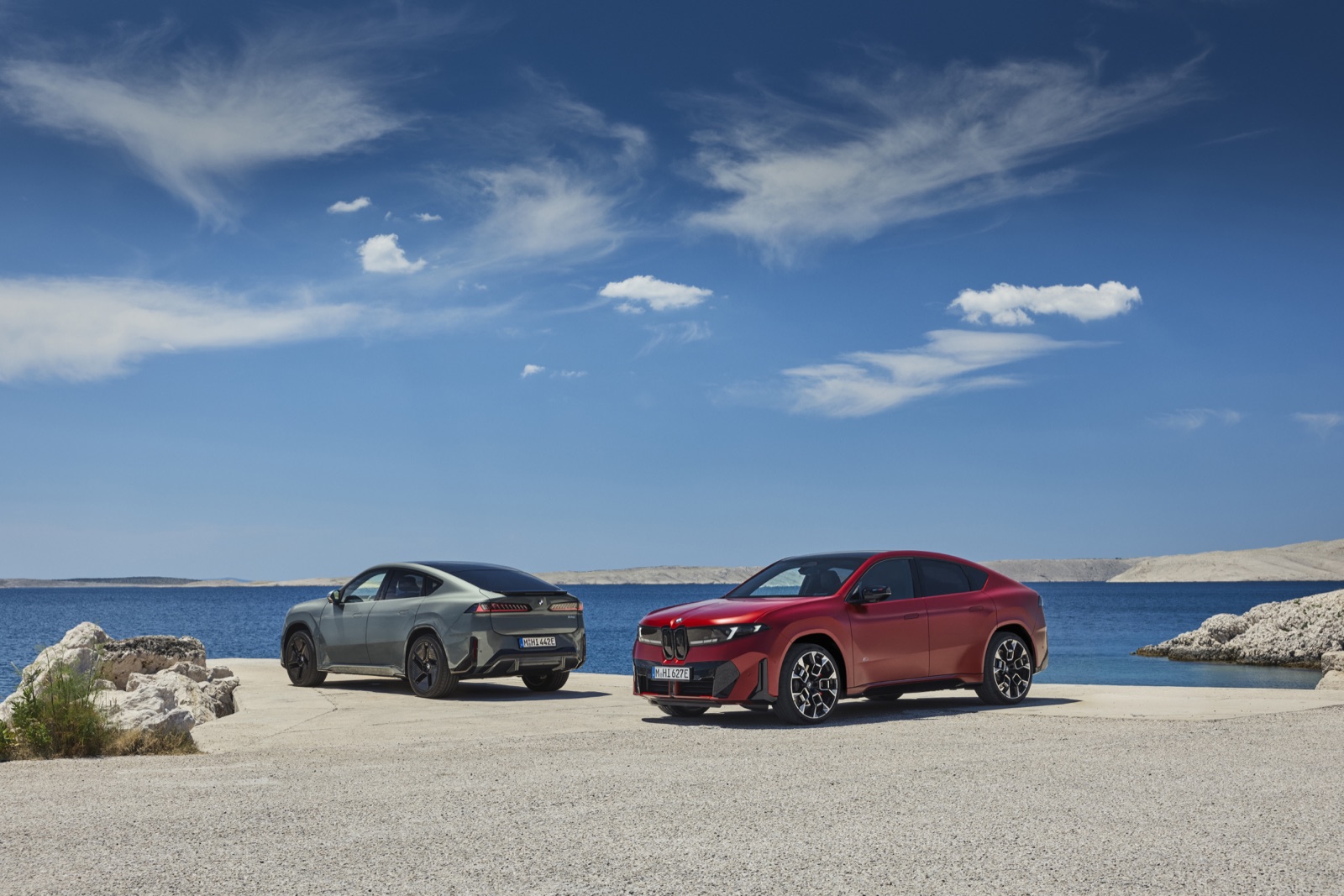 Dve verzie nového BMW iX4 na pobrežnej ceste, červená a sivá.